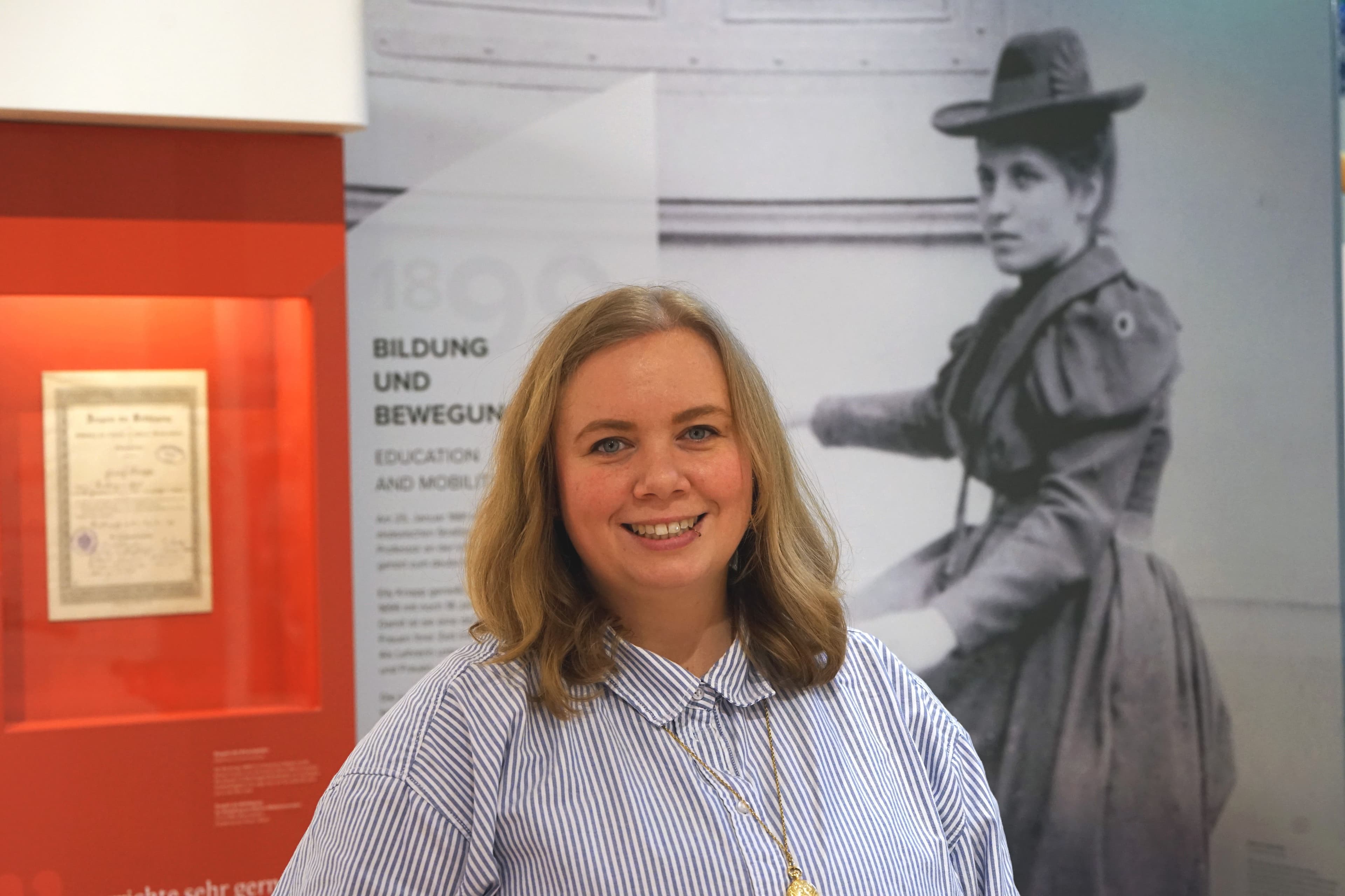 Porträtfoto einer Frau mit schulterlangen blonden Haaren und einem blau-weiß gestreiften Hemd. Sie lächelt freundlich in die Kamera. Im Hintergrund ist eine Ausstellungstafel mit dem Titel „Bildung und Bewegung" sowie ein historisches Schwarz-Weiß-Foto einer Frau in Kleidung des späten 19. Jahrhunderts zu sehen.